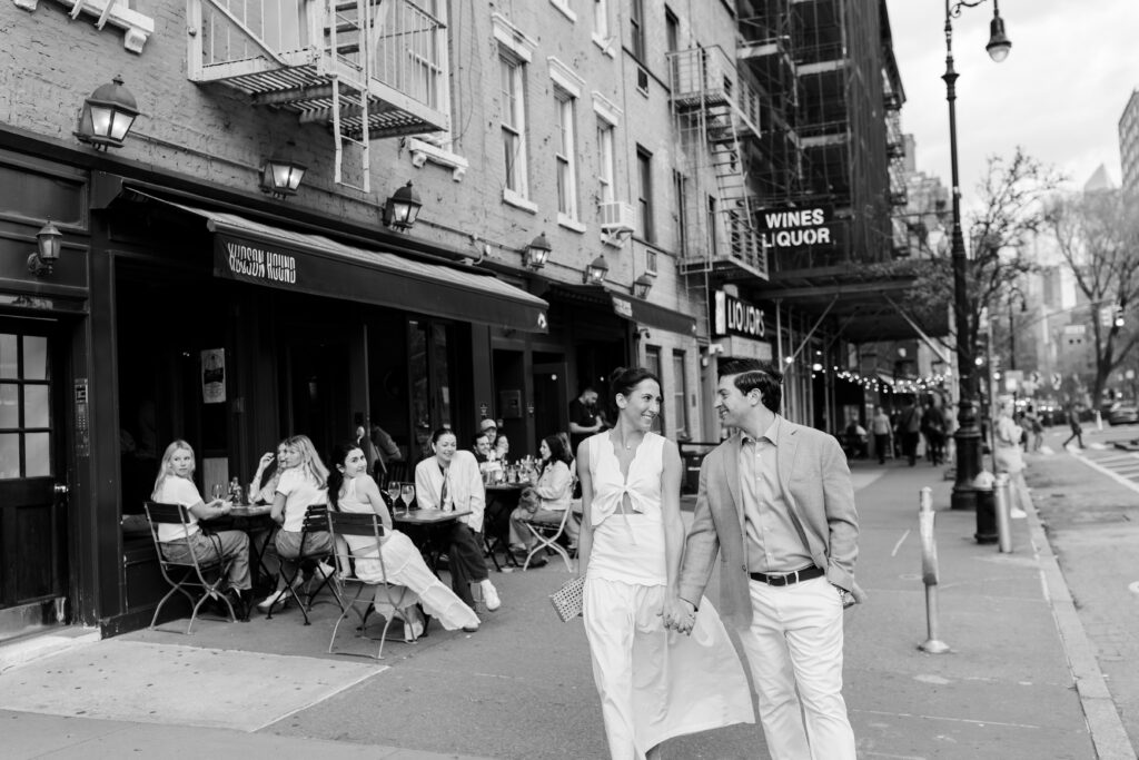 couple smiling and holding hands walking down West Village in front of Hudson Hound for their engagement photos