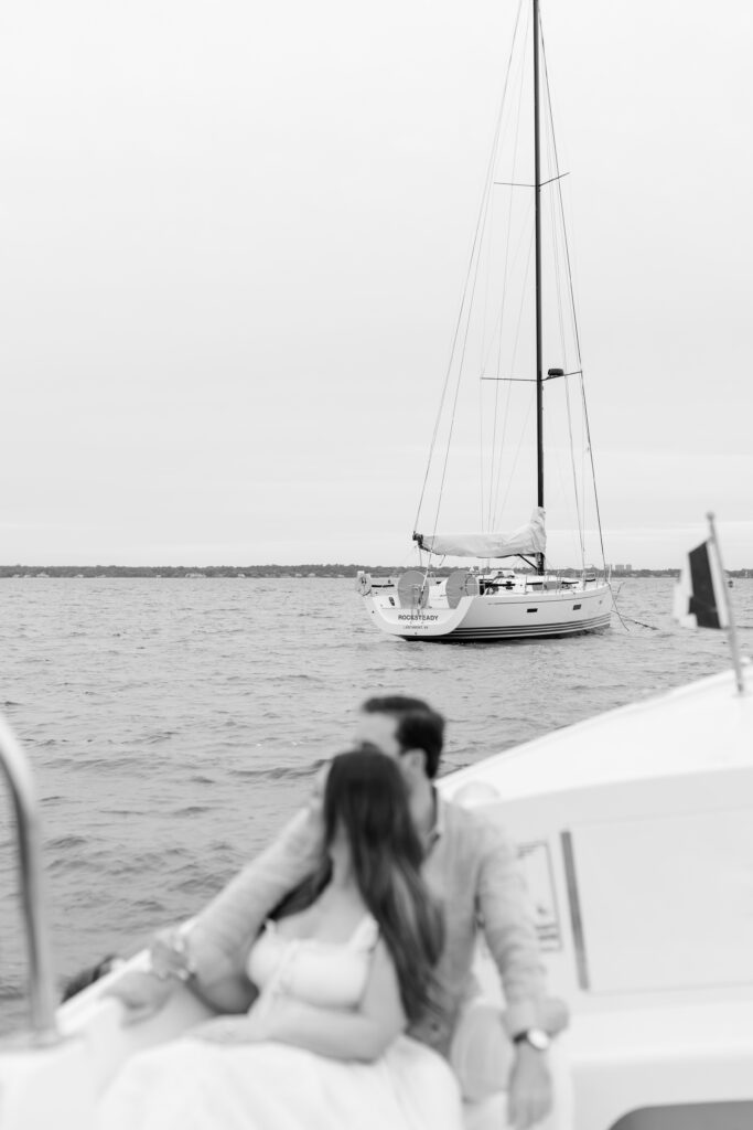 couple sitting on the sailboat during their engagement session at Larchmont Yacht Club with a passing boat in the background