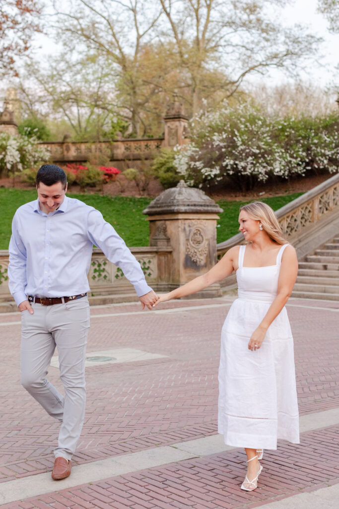 couple laughing and walking while holding hands in by the stairs of the Bethesda Terrace in Central Park during their golden hour engagement session