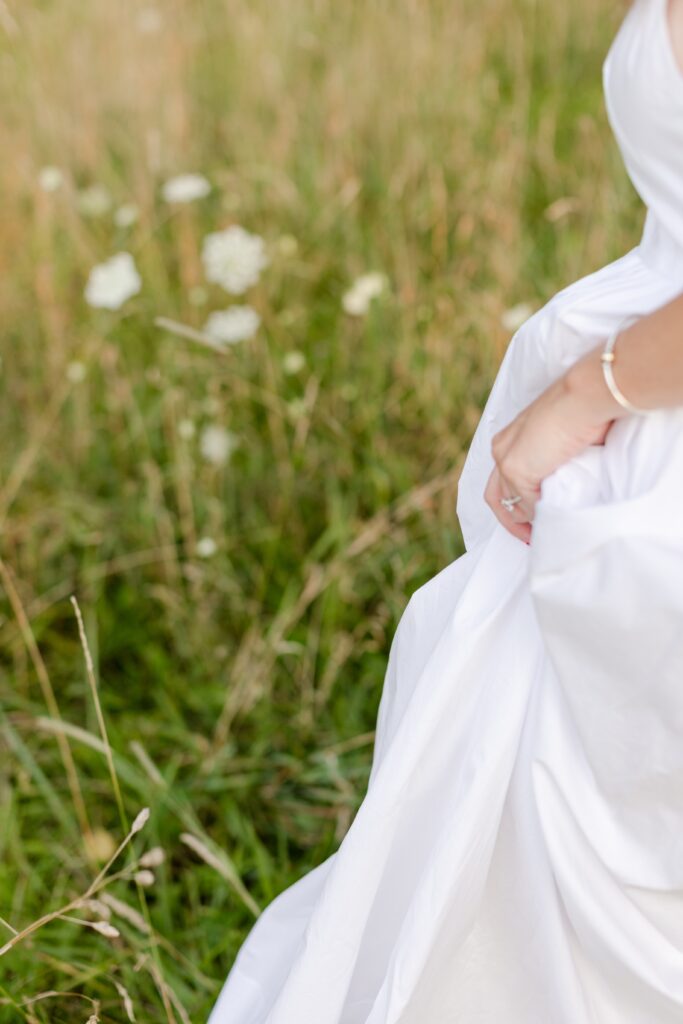 woman holding her white dress while walking through the field for an engagement session at Rockefeller State Park