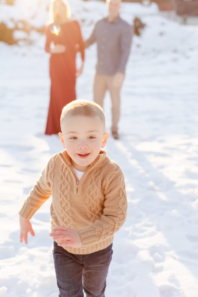 boy running towards camera in the snow with parents in the background during engagement session at Harts Brook Park and Preserve