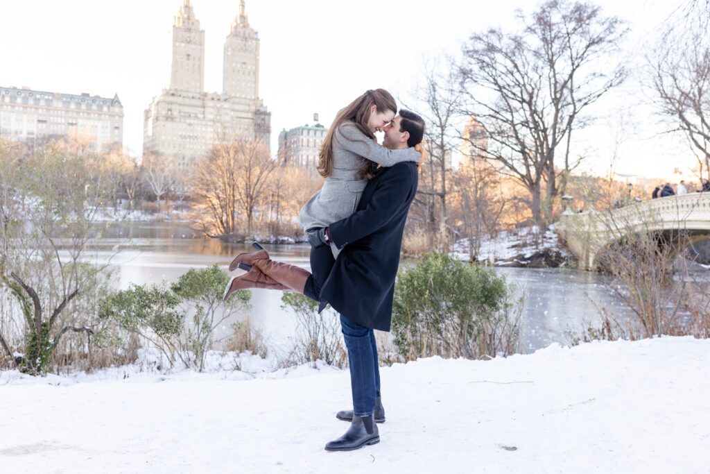 guy holding fiance in snowy Central Park with El Dorado in the background during winter engagement session